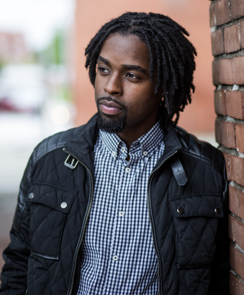 Portrait of a man in a dress shirt and black jacket standing against a brick wall.